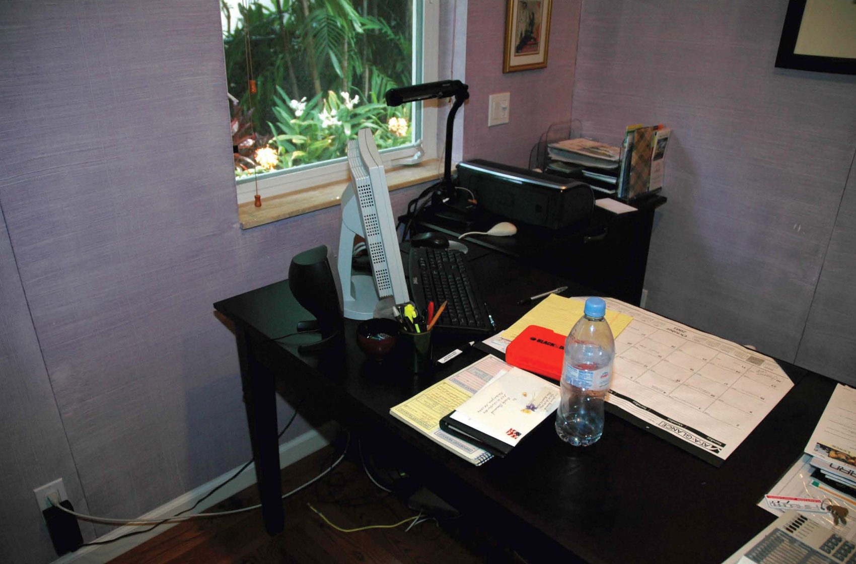 Schreibtisch mit Monitor, Tastatur, Lampe und Papieren in einem abgedunkelten Arbeitszimmer